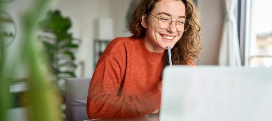 Happy,Young,Woman,Using,Laptop,Sitting,At,Desk,Writing,Notes